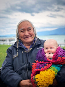 In the foreground, an Elder dressed in a blue fleece with white hair in two braids holds a small baby wrapped in a rainbow colored quilted blanked. The coast line, a boardwalk, and mountains are blurred in the background.